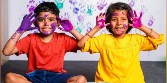 Image of two kids with holi colours on their hands and big smiles on their faces. They are holding their hands with their palms facing the user to make it seem like they have big ears.